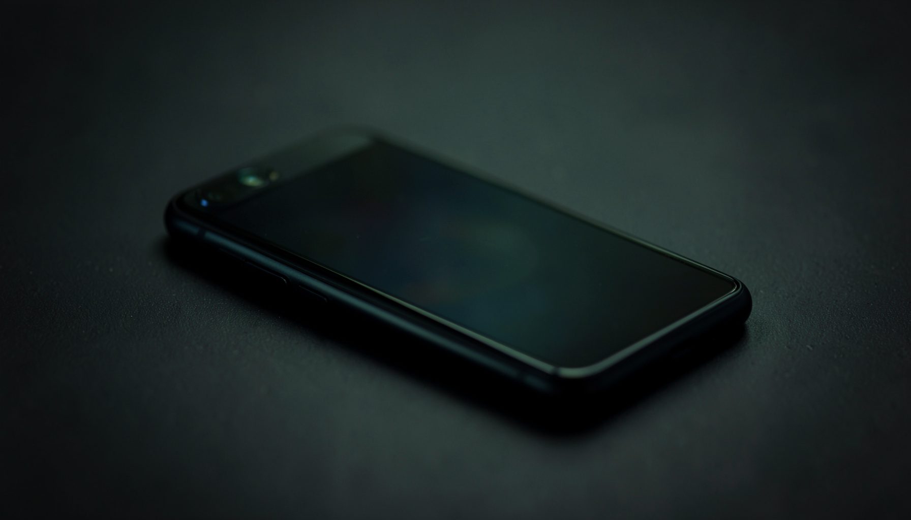 Gros plan sur un smartphone aux reflets sombres posé sur une surface mate, ambiance tamisée vert foncé et noir